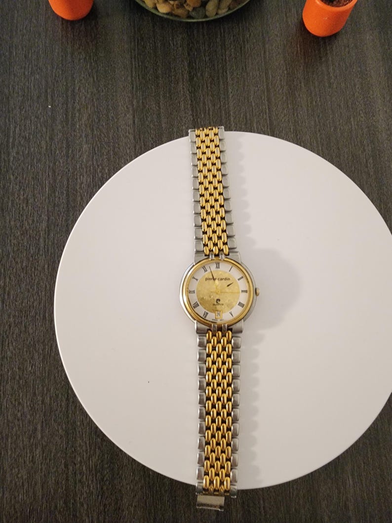May include: A gold and silver-toned Pierre Cardin wristwatch. The watch has a gold face with black markers and hands. The band is a combination of gold and silver links. The watch is displayed on a white surface.