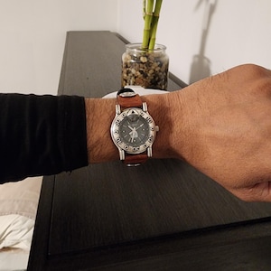 May include: A wristwatch with a brown leather band and a silver-colored face. The watch face has black numerals and a unique design. The watch is on a person's wrist.