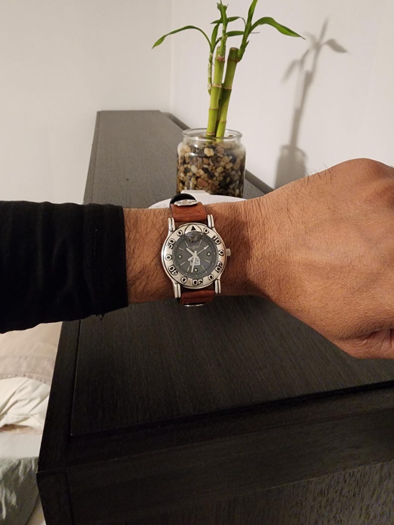 May include: A wristwatch with a brown leather band and a silver-colored face. The watch face has black numerals and a unique design. The watch is on a person's wrist.