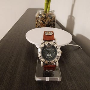 May include: A silver-toned wristwatch with a brown leather band is displayed on a clear acrylic stand. The watch face is dark with silver accents and the brand name "BOY LONDON". The watch is set against a neutral background.