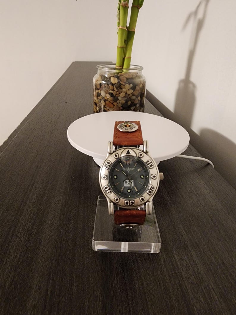 May include: A silver-toned wristwatch with a brown leather band is displayed on a clear acrylic stand. The watch face is dark with silver accents and the brand name "BOY LONDON". The watch is set against a neutral background.