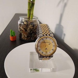 May include: A Pierre Cardin watch with a gold-toned face and silver and gold-toned band. The watch has Roman numeral hour markers and a date display. The watch is displayed on a clear acrylic stand.