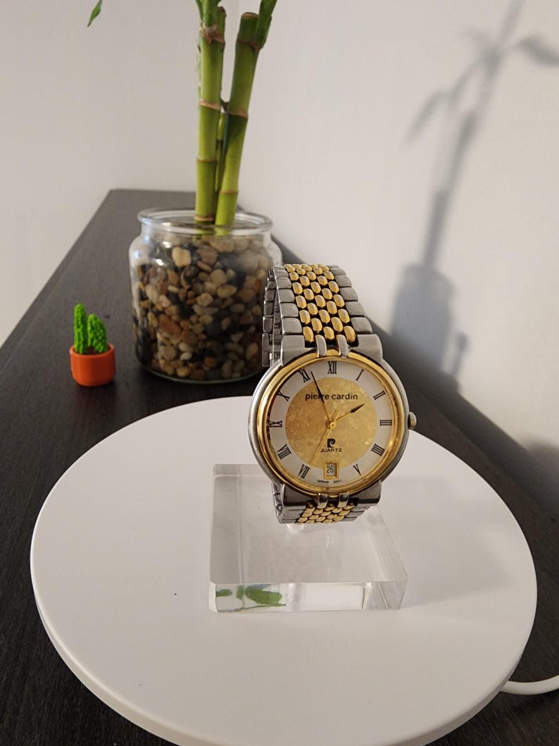 May include: A Pierre Cardin watch with a gold-toned face and silver and gold-toned band. The watch has Roman numeral hour markers and a date display. The watch is displayed on a clear acrylic stand.