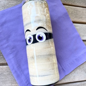 May include: A white and black mummy-themed tumbler with purple eyes. The tumbler is on a purple napkin.