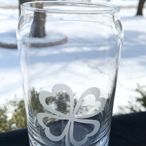 May include: A clear glass can with a four-leaf clover etched on the side. The can is sitting on a black surface.