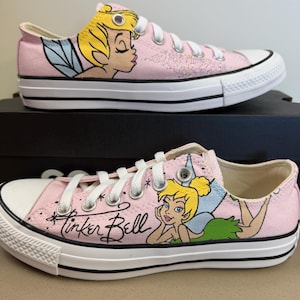 May include: A pair of light pink low-top canvas sneakers featuring illustrations of Tinker Bell. The shoes have white laces, white rubber toe caps, and white soles with black stripes. One shoe shows Tinker Bell in profile, while the other displays her reclining with the script text Tinker Bell written in black cursive.