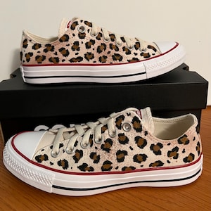 May include: A pair of low-top Converse sneakers with a leopard print design. The sneakers are white with brown and black spots. The sneakers are on a wooden surface.