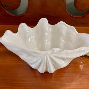 May include: A white ceramic bowl shaped like a giant clam shell. The bowl features a glossy finish with a textured, undulating rim and a fan-like pleated base. The interior is smooth and deep, mimicking the natural form of a mollusk shell.
