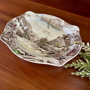 May include: An oval-shaped ceramic serving dish with a detailed landscape scene in brown, white, and green. The dish features a scalloped edge and a vintage aesthetic, perfect for displaying or serving food.
