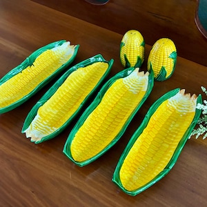 May include: A set of four ceramic corn-shaped serving dishes and two matching corn-shaped salt and pepper shakers. Each dish features a bright yellow textured cob center with green husk edges. The shakers are yellow with green base details.