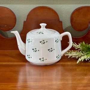 May include: A white ceramic teapot featuring a repeating pattern of small green shamrocks across the body and lid. The teapot has a curved handle, a tapered spout, and a matching removable lid with a small knob handle on top.