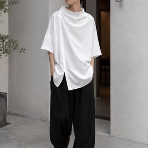 May include: A white short-sleeved shirt with a high collar and an asymmetrical button placket running down the front, paired with loose-fitting black trousers and black open-toe sandals. The shirt features a smooth fabric and a relaxed fit.
