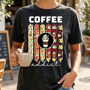 Op de afbeelding: Een donkergrijs t-shirt met het woord "COFFEE" in witte blokletters. Daaronder verticale strepen met koffie-thema illustraties. Het woord "addict" staat eronder in cursief. De persoon houdt een koffiekopje vast.