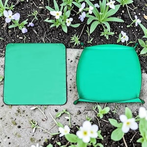 Puede incluir: Dos rodilleras verdes para jardín. Una es cuadrada, la otra es redondeada con asas. Las almohadillas están colocadas sobre una superficie de hormigón con tierra y pequeñas flores blancas y moradas en el fondo. Accesorios de jardinería.