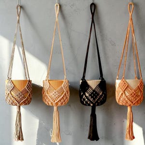 Puede incluir: Cuatro colgadores de plantas de macramé en varios colores: beige, melocotón, negro y naranja. Cada uno presenta un diseño tejido alrededor de una maceta de color claro, con una larga borla en la parte inferior. Los colgadores están suspendidos de ganchos metálicos.