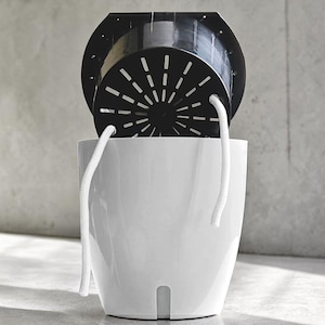 Op de afbeelding: Een witte plastic plantenpot met een zwarte binnenpot. De zwarte pot heeft een geperforeerd ontwerp en wordt door twee witte koorden boven de witte pot gehangen. De pot is ontworpen voor binnen of buiten.