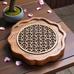 May include: A decorative wooden coaster with a carved, geometric pattern in the center. The coaster has a scalloped edge and a warm, brown finish. The coaster is on a wooden table with a small bonsai tree and scattered pink flower petals.
