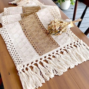 May include: A cream and brown striped woven table runner. The runner has a textured weave and a fringe trim along the bottom edge. A small bunch of dried flowers is placed on the runner.
