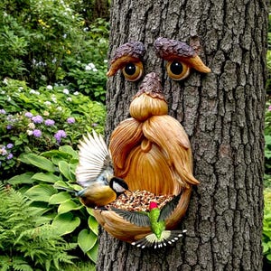 Puede incluir: Un comedero para pájaros de madera, tallado para parecer una cara, montado en un tronco de árbol. El comedero tiene un diseño similar a una barba y ojos grandes. Se muestran dos pájaros alimentándose del cuenco. El comedero está rodeado de exuberante follaje verde.