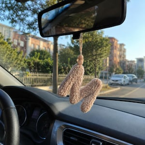 May include: A beige, crocheted car charm in the shape of the letter "M" hangs from a rearview mirror. The charm is suspended by a natural-colored string and wooden bead. The car's dashboard and steering wheel are visible.