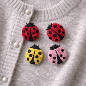 Op de afbeelding: Close-up van een grijze cardigan versierd met vier gehaakte lieveheersbeestjes applicaties. Twee zijn rood met zwarte stippen, een is geel met zwarte stippen en een is roze met zwarte stippen. De cardigan heeft parelknopen.