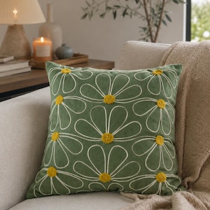 Op de afbeelding: Een decoratief groen sierkussen met een madeliefjespatroon. Het kussen heeft witte contouren en gele centra op het madeliefjesontwerp. Het ligt op een beige bank met een beige deken.