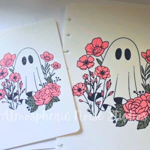 May include: Two white planner inserts featuring a black-outlined ghost surrounded by coral-pink flowers and dark green leaves. The ghost has black-filled eyes. The text Atmospheric Noise Studios is printed in white script at the bottom center.