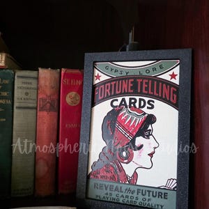 May include: A framed rectangular sign featuring the text Gypsy Lore Fortune Telling Cards and Reveal the Future. The graphic depicts a profile illustration of a person wearing a headpiece, rendered in red, black, and cream colors on a white background.