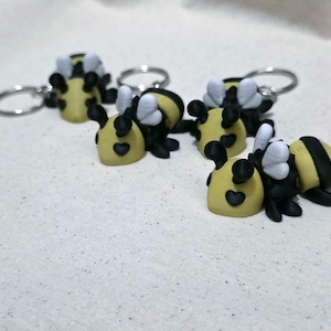 May include: Four bee-shaped keychains featuring yellow and black striped bodies, small white wings, and a heart-shaped detail on the face. Each bee is attached to a silver-toned metal split ring, arranged in a cluster on a light-colored fabric surface.