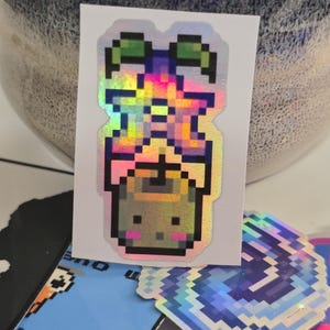 May include: A holographic sticker featuring a pixelated plant in a pot with a smiling face. The sticker has a rainbow sheen and is surrounded by other stickers with pixelated designs.