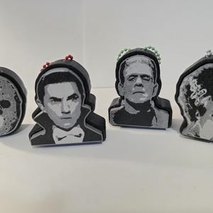 May include: Four black foam keychains featuring gray and white graphic illustrations of horror characters, including a hockey mask, a vampire, Frankenstein's monster, and the Bride of Frankenstein, each attached to a small colored ball chain.