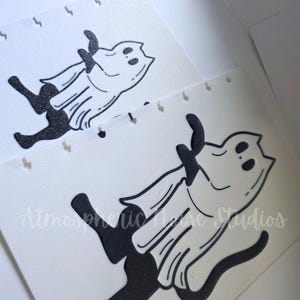 May include: Two white disc-bound planner inserts featuring a black glittery graphic of a cat wearing a ghost costume. The cat is depicted in a running pose with a long tail. The text Atmospheric House Studios is printed in a light script font.