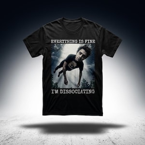 Op de afbeelding: Zwart t-shirt met de tekst "EVERYTHING IS FINE" boven een afbeelding van een vallende persoon en de tekst "I'M DISSOCIATING" eronder. De afbeelding heeft een surrealistische, dromerige kwaliteit met een donkere achtergrond.