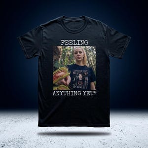 Op de afbeelding: Zwart t-shirt met de tekst "FEELING ANYTHING YET?" boven en onder een foto van een persoon die een Venus vliegenval vasthoudt. De persoon draagt een shirt met de tekst "LOGGED OUT OF REALITY".