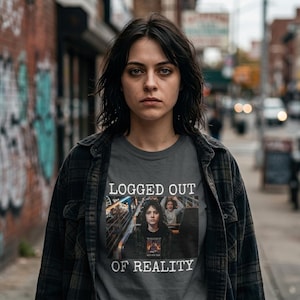 Op de afbeelding: Een grijs t-shirt met de woorden "LOGGED OUT OF REALITY" in het wit gedrukt. Het shirt heeft een afbeelding van een persoon in een attractie. De persoon draagt een zwart-grijs geruit overhemd.
