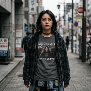 Op de afbeelding: Een donkergrijs t-shirt met de tekst "LOGGED OUT OF REALITY" en een afbeelding van een persoon. De persoon draagt een zwart-grijs geruit jack en een blauwe spijkerbroek. De achtergrond is een stadsstraat.