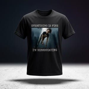 Op de afbeelding: Zwart t-shirt met de witte tekst "EVERYTHING IS FINE I'M DISSOCIATING". De afbeelding toont een persoon die in een donker, surrealistisch landschap valt.