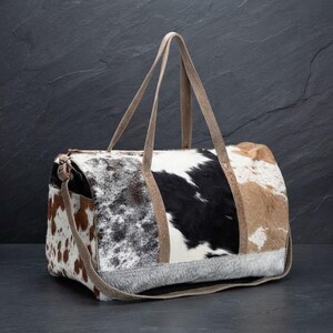 Puede incluir: Un bolso de viaje hecho de piel de vaca con un diseño de retazos de pelo marrón, negro, blanco y gris. El bolso incluye dos asas superiores largas y un panel base de pelo gris texturizado, con forma rectangular y cierre de cremallera.