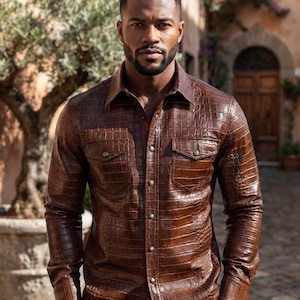 Peut inclure: Une chemise à manches longues boutonnée d'une couleur marron riche avec un motif texturé en peau de crocodile. La chemise présente un col classique, deux poches de poitrine à rabats boutonnés et une patte de boutonnage centrale avec des boutons métalliques.