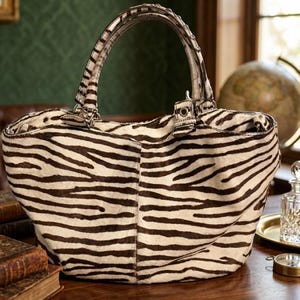 May include: A handbag featuring a brown and white zebra stripe pattern with a textured hair-on-hide finish. The bag includes a single arched handle with metallic stud accents, a central seam, and a structured opening with side hardware attachments.