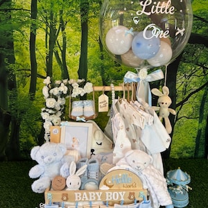 May include: A baby gift basket with a variety of items, including clothing, socks, stuffed animals, and a framed picture. The basket is decorated with blue and white accents and the words "Baby Boy". A large clear balloon with the text "Hello Little One" floats above the basket.