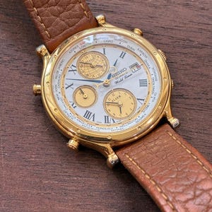 May include: A gold-tone Seiko World Timer quartz watch with a white dial, Roman numerals, and three sub-dials. The watch features a world time bezel listing global cities and is attached to a textured brown leather strap with visible stitching.