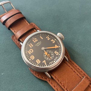 Könnte beinhalten: Eine Timex Midget Armbanduhr mit rundem silberfarbenem Gehäuse, dunkelgrauem Zifferblatt und orangefarbenen arabischen Ziffern. Die Uhr hat orangefarbene Zeiger, ein kleines Sekunden-Hilfszifferblatt und ein breites braunes Lederarmband mit Silberschnalle.