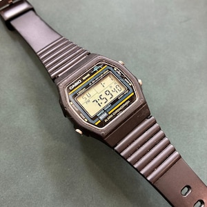 May include: A black digital wristwatch with a ribbed resin strap. The rectangular face features a gray LCD screen displaying the time 7:59:40, the date, and the text CASIO, Lithium, Water Resist 50M, and Alarm Chronograph with yellow and blue accents.