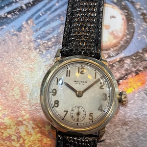 Op de afbeelding: Een vintage Waltham horloge met een zilveren wijzerplaat, goudkleurige kast en zwarte leren band. De wijzerplaat toont de tekst "WALTHAM MOVEMENT" en heeft Arabische cijfers. De band is ongeveer 2 cm breed.
