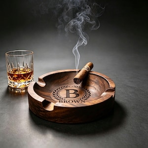 May include: A round dark wood ashtray featuring a central engraved laurel wreath design with the letter B and the name BROWN below it. A lit cigar rests across the rim with visible smoke rising, accompanied by a glass of amber-colored liquid.