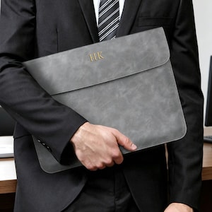 May include: A grey document sleeve with a smooth, matte finish and a flap closure. The front of the sleeve features the gold-coloured embossed initials HK centred near the top edge. The sleeve has rounded corners and a slim, rectangular profile.