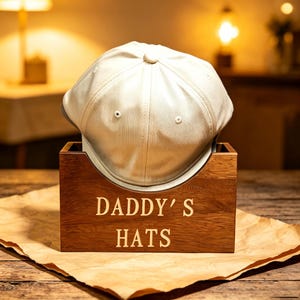 May include: A cream-colored baseball cap resting inside a rectangular wooden storage box. The front of the box features the engraved text DADDY'S HATS in a clean, sans-serif font. The cap has a structured crown with a central button detail.