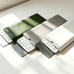 以下が含まれることがあります： オリーブグリーン、グレー、ホワイトの長方形のキーボードスリーブ3点。各スリーブは滑らかな仕上げで、キーボード用の切り込みがあり、上部のフラップにはゴールドでAHのイニシャルが刻印されています。白いキーボードがそれぞれ部分的に挿入されています。