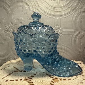 May include: A light blue glass candy dish shaped like a high-heeled shoe. The dish has a hobnail pattern and a lid with a small knob. The shoe-shaped dish is sitting on a cream-colored, crocheted doily.